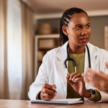 A hardworking African American female doctor sitting at the wooden table with notes and talking with a patient during a home visit.